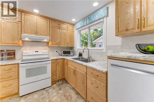 Wood-finish cabinetry, light-toned countertops, tile backsplash, white appliances, and a double basin sink - 422 Grangewood Drive, Waterloo, ON 