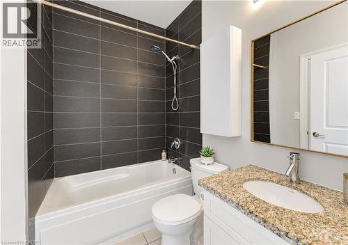 Bathroom featuring a built-in bathtub with dark tiled surround, white toilet, vanity with a light speckled countertop, and a rectangular mirror with a gold-tone frame - 25 Concession Street Unit# 110, Cambridge, ON 