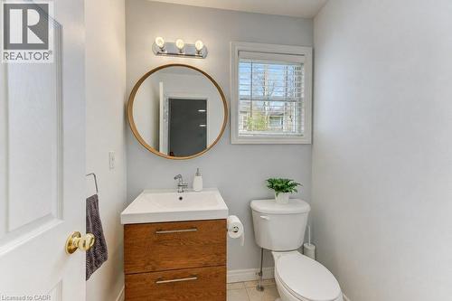 White paneled door, wood-finish vanity with chrome hardware, circular framed mirror, three-light vanity fixture, and a window with blinds - 88 Decaro Crescent, Cambridge, ON 