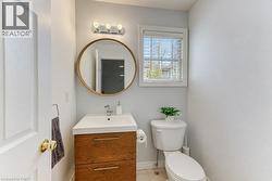 White paneled door, wood-finish vanity with chrome hardware, circular framed mirror, three-light vanity fixture, and a window with blinds - 