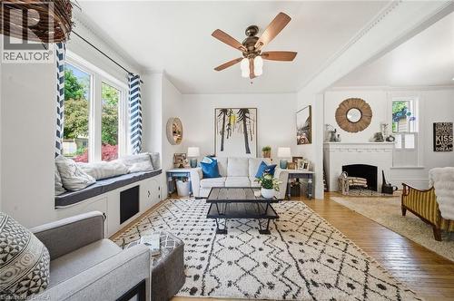 Living area featuring wood-finish flooring, a white brick fireplace, a built-in window seat with integrated storage, a ceiling fan with light fixture, and crown molding - 62 East Avenue, Brantford, ON 