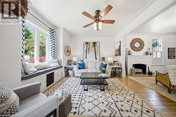 Living area featuring wood-finish flooring, a white brick fireplace, a built-in window seat with integrated storage, a ceiling fan with light fixture, and crown molding - 