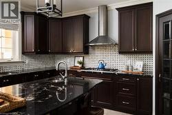 Dark wood cabinetry with crown molding, black stone countertops, a tile backsplash with black accents, a stainless steel range hood, and a built-in kitchen sink - 