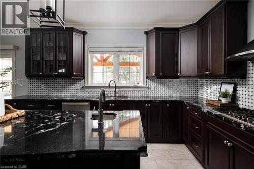 Dark wood cabinetry with glass-front uppers, stone countertops, stainless steel appliances, a tiled backsplash, and light-toned tile flooring - 1505 Milburough Line, Carlisle, ON 