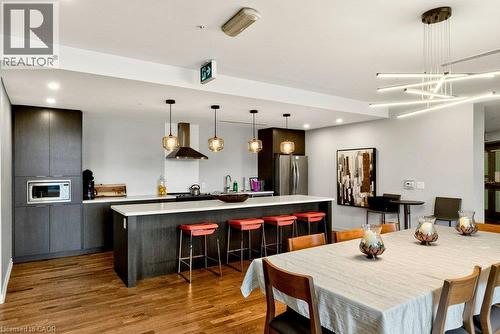 Open-concept kitchen and dining area featuring rich wood-finish flooring, dark wood-finish cabinetry, a large kitchen island with white countertop, and a modern linear chandelier - 457 Plains Road Unit# 225, Burlington, ON 