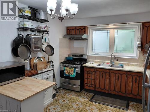 Kitchen featuring wood-finish cabinetry, a double basin stainless steel sink, a gas range with a range hood, and patterned floor tiling - 244 Guildwood Drive, Hamilton, ON 