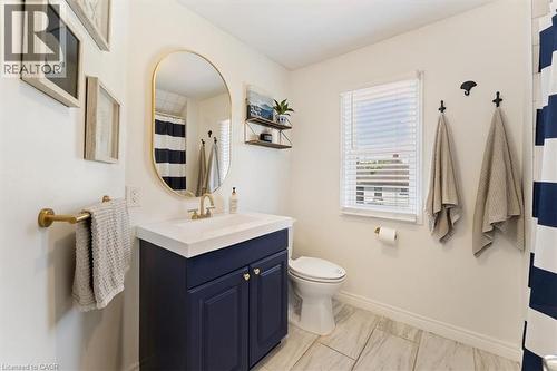 Bathroom featuring a navy blue vanity with a white countertop and gold-finish hardware, a gold-framed oval mirror, a white toilet, and light-toned tile flooring - 17 Craigmiller Avenue, Hamilton, ON 
