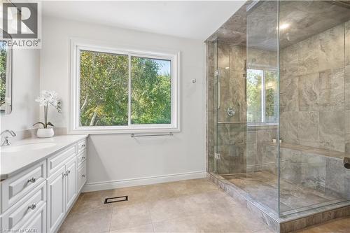 Spacious bathroom featuring a glass-enclosed shower with a built-in bench, a window, a vanity with a white countertop and double-door cabinetry, and neutral floor tiling - 4159 Spruce Avenue, Burlington, ON 
