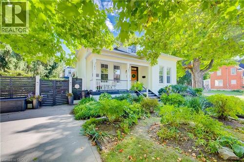 Light-colored exterior with a front porch, mature trees, landscaped garden beds, and a dark-toned fence with gate - 59 Allen Street W, Waterloo, ON 