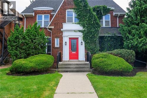 Brick facade residence featuring a prominent red entry door, white portico, symmetrical shrubbery, a concrete walkway, and a landscaped front yard - 414 Main Street E, Hamilton, ON 