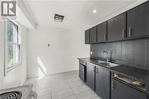Black cabinetry with gold-tone hardware, dark countertop, dual basin stainless steel sink, subway tile backsplash, and light-toned tile flooring - 70 Fiddlers Green Road Unit# 74, London, ON 