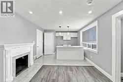 Open-concept interior featuring wood-finish flooring, a white decorative fireplace mantel with tile surround, a kitchen island with a neutral countertop, contemporary pendant lighting, and crisp white - 