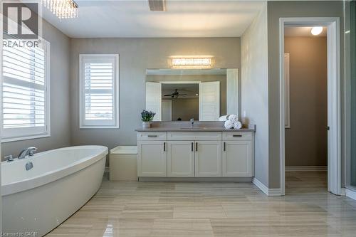 Freestanding soaking tub, white plantation shutters, dual-sink vanity with light-toned countertop, large wall-mounted mirror, and tile flooring - 455 Equestrian Way, Cambridge, ON 