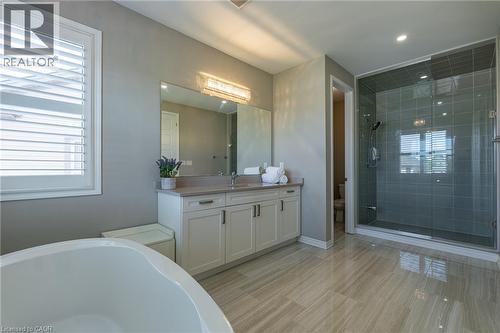 Spacious bathroom featuring a freestanding tub, double vanity with stone countertop, glass-enclosed shower with tiled walls, wood-finish tile flooring, and window with plantation shutters - 455 Equestrian Way, Cambridge, ON 