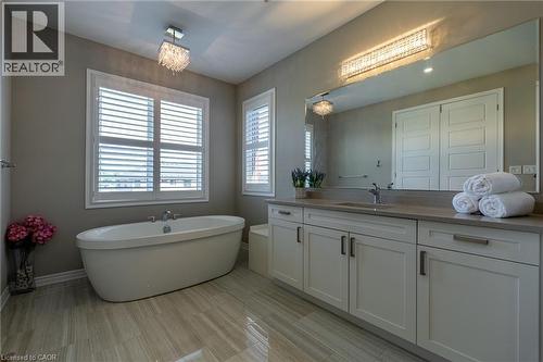 Freestanding oval soaking tub, dual-pane windows with plantation shutters, wood-finish tile flooring, extended vanity with light-toned countertop, and white shaker-style cabinetry - 455 Equestrian Way, Cambridge, ON 