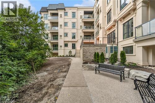 Contemporary building exterior with multiple balconies featuring glass railings, a stone retaining wall, and an exterior staircase - 2 Arbourvale Common Unit# 209, St. Catharines, ON - Outdoor With Balcony With Facade
