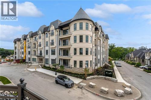 Multi-story residential building with a stone and stucco facade, featuring multiple balconies with iron railings, a prominent turret with a conical roof, and a grand entrance with stone stairs - 2 Arbourvale Common Unit# 209, St. Catharines, ON - Outdoor With Facade