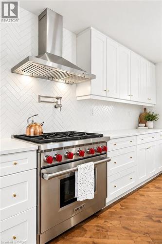 Stainless steel range with red control knobs, white shaker-style cabinetry, white countertops, wood-finish flooring, and a white herringbone tile backsplash with a pot filler - 9 Midway Street, Hamilton, ON 