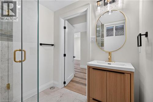 Glass-enclosed shower with gold-finish hardware, light wood-finish vanity with integrated sink, gold-finish oval mirror, and three-light vanity fixture - 3028 Eva Drive, Burlington, ON 
