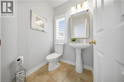 Pedestal sink with chrome faucet, wall-mounted mirror, window with white louvered blinds, tile flooring, and a two-piece toilet - 3352 Hiscott Avenue, Burlington, ON 