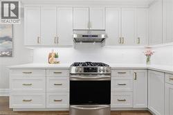 White shaker cabinetry with gold-tone hardware, stainless steel gas range, coordinating range hood, white subway tile backsplash, and light-toned countertops - 