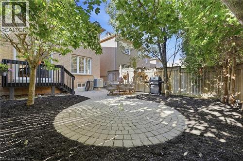Circular paver patio, wood deck with black railing, mature trees, wood fence, and brick exterior - 2409 Postmaster Drive, Oakville, ON 