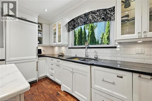 White cabinetry with glass-front uppers, dark solid surface countertops, a subway tile backsplash, panel-front refrigerator, and wood-finish flooring - 2209 Ghent Avenue, Burlington, ON 