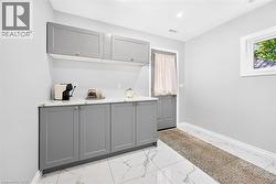 Gray shaker-style cabinetry with gold-tone hardware, white countertop, white tile flooring with gray veining, recessed ceiling lighting, and a white-framed window - 
