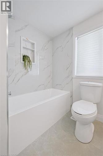 Bathroom featuring a white bathtub with a built-in shower niche, marble-look tile surround, a window with privacy blinds, and light-toned tile flooring - 299 Dale Crescent, Waterloo, ON 