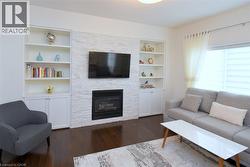 Bright, open living room featuring a white brick fireplace with built-in shelving and cabinetry, wall-mounted flat-screen television, wood-finish flooring, and abundant natural light from multiple win - 