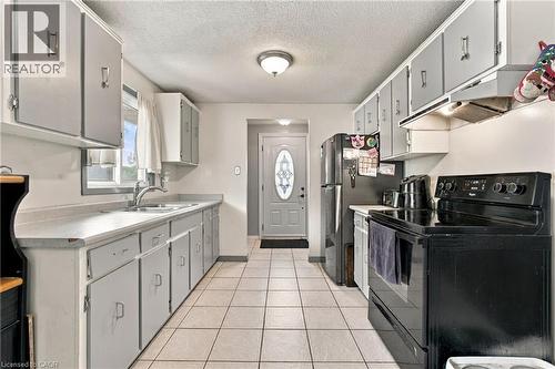 Galley kitchen featuring tile flooring, white cabinetry with gray panels, a double basin sink, and a front entry door with decorative glass - 135 Cedarhill Crescent, Kitchener, ON 