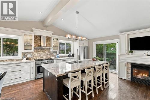 Vaulted ceiling with exposed beams, white perimeter cabinetry with glass-front accents, stainless steel range with matching hood, large kitchen island with integrated sink, and a built-in fireplace wi - 617 Scenic Drive, Hamilton, ON 
