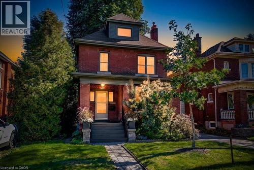 Red brick exterior with a gray-shingled roof, featuring a covered porch with stone-clad pillars, a dormer window, and a paved walkway - 43 Rosslyn Avenue S, Hamilton, ON 