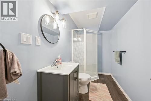 Vanity with integrated sink and chrome faucet, round wall-mounted mirror, three-light sconce, enclosed shower, and wood-finish flooring - 765 Angler Way, Waterloo, ON - Indoor Photo Showing Bathroom