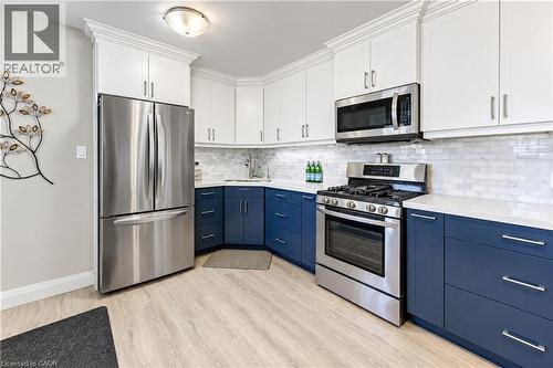 Kitchen featuring two-tone cabinetry with white upper and blue lower cabinets, stainless steel appliances, light-toned wood-finish flooring, white subway tile backsplash, and light-toned countertops - 87 Bond Street, Kitchener, ON 