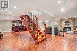 Open-concept living space featuring wood-finish flooring, a central staircase with wooden treads and wrought-iron balusters, a built-in wet bar with cabinetry, and a stone-clad fireplace with a hearth - 