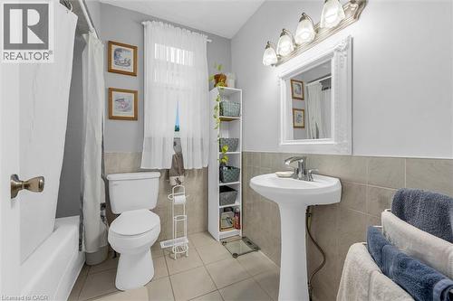 Full bathroom featuring a pedestal sink, a bathtub-shower combination, a wall-mounted mirror with an integrated light fixture, and tile flooring with matching wainscot tiling - 143 Walter Avenue S, Hamilton, ON 