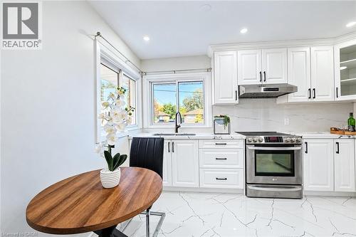 Kitchen featuring white Shaker-style cabinetry, stainless steel range with overhead vent hood, stone-finish countertops, full-height stone-finish backsplash, and light-toned tile flooring with veining - 46 Traynor Avenue, Kitchener, ON 
