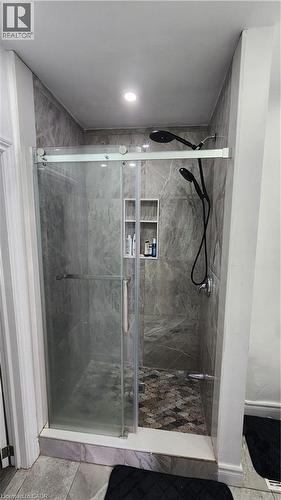 Walk-in shower featuring floor-to-ceiling stone-finish tile, sliding glass door enclosure, pebble-stone flooring, built-in recessed shelving, and a dual-head matte black rainfall shower system - 1394 Cannon Street E, Hamilton, ON 