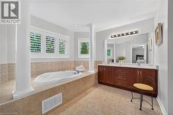 Jetted soaking tub with tile surround, double-sink wood vanity, large wall-mounted mirror, decorative structural columns, and plantation shutters - 