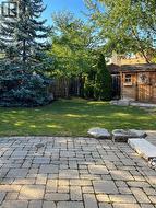 Backyard featuring a stone paver patio, natural stone accents, mature evergreen trees, and a detached wood-clad storage structure with a shingle roof - 