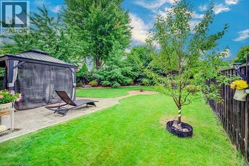Backyard featuring a paved patio, covered structure with a metal roof, circular stone fire pit area, manicured lawn, and mature trees enclosed by a wooden fence - 521 Snowdrop Court, Kitchener, ON 