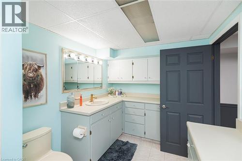 Bathroom featuring a vanity with integrated sink, brass faucet, expansive wall-mounted mirror with multi-bulb lighting, built-in upper and lower cabinetry, and a panelled door - 43 Alderney Avenue, Hamilton, ON 
