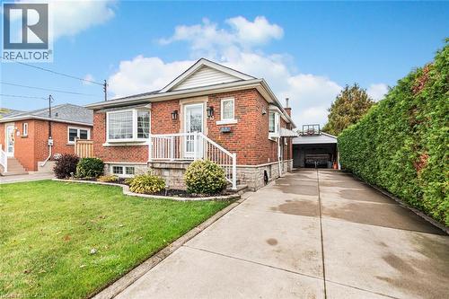 Red brick exterior featuring a stone-faced foundation, a concrete driveway, a detached garage, and a raised front porch with white railing - 43 Alderney Avenue, Hamilton, ON 