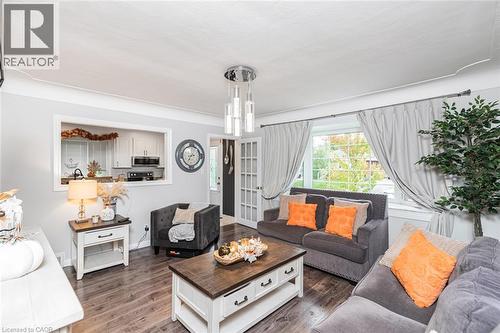 Living area featuring wood-finish flooring, a multi-light modern pendant fixture, a picture window, and a built-in kitchen pass-through window with a surrounding frame - 43 Alderney Avenue, Hamilton, ON 