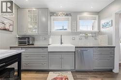 Kitchen featuring gray shaker-style cabinetry, a white farmhouse sink with a gooseneck faucet, light-toned stone-finish countertops, glass-front upper cabinet detailing, and wood-finish flooring - 
