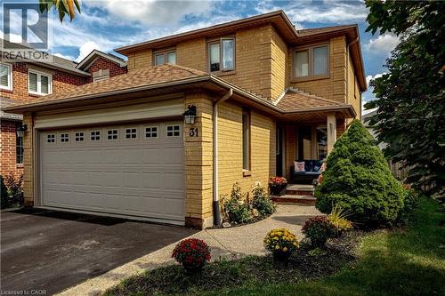 Brick exterior with a covered entry, attached garage with decorative windows, paved driveway, and established front landscaping - 31 Grindstone Way, Hamilton, ON 