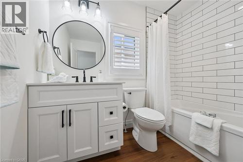 White vanity with black hardware, round mirror, dark wood-finish flooring, subway tile bath surround, and white plantation shutters - 2294 Seton Crescent, Burlington, ON 