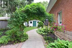 Arched garden trellis, concrete pathway, red brick exterior, landscaped garden beds, and a white storage shed with a green door - 