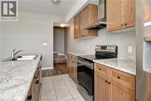 Galley kitchen featuring wood-finish cabinetry, granite-style countertops, stainless steel electric range with hood ventilation, and tile flooring - 102 Wheat Lane, Kitchener, ON 